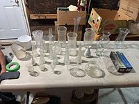 Wide view of 19 assorted glass and plastic vases in varied styles and sizes on table.