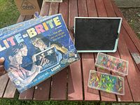 Vintage Lite-Brite box, pegboard/lightbox and two cases of translucent pegs laid out on wooden surface.