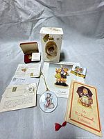 Group shot of all Hummel/Goebel items: egg ornament with box, round ornament, rectangular pin, gold medallion with case, booklets and paperwork