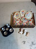 Overall lot showing tea set in patterned box, additional cups and saucers on black insert, and three porcelain doll heads