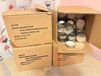 Three cartons, one open showing stacked silver screw-top tins with packing