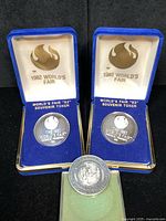 Two 1982 World's Fair silver souvenir tokens in blue velvet boxes with 'World's Fair '82 Souvenir Token' inscription and one Newton MA Centennial silver coin in a green felt box.