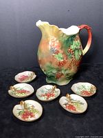 Porcelain jug and six grape leaf-shaped dishes displayed against a dark background, showing frontal view with jug on right and dishes arranged in front