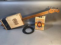 Full view of 3-string cigar box guitar with attached tuning head, amplifier made from Indian Tabac Cigar Co. box, and cable coiled beside them.