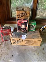 Photo showing all items in the set: original boxed lantern, camp stove box, Coleman Silk-Lite mantles, and gallon can of Coleman Fuel on a dusty floor.