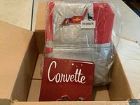 Car cover folded inside clear plastic packaging showing red and silver colors and embroidery with Chevrolet and Corvette logos, placed inside an open cardboard box with the book titled 'Corvette' in front.