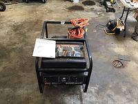 Full view of generator on garage floor with rolled orange extension cord and manual on top
