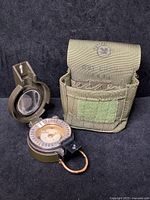 Photo showing Israeli military compass with oil-filled dial placed open next to olive green ammo pouch waist bag with clip.