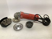 Milwaukee 5 inch sander-grinder shown with four sanding and grinding attachments arranged on a white surface.