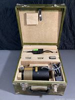 Open green storage case showing the black cylindrical polariscope unit with perforated gold guard, cable attachments, and compartments