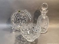 Three Waterford Crystal pieces on a neutral grey background including a Celtic knot patterned plate, a footed bowl with star cut design, and a decanter with faceted stopper.