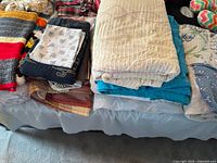 Overview of various folded quilts, blankets, and pillowcases stacked on a bed with crocheted and patterned linens visible.