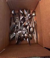 Top view of a cardboard box containing a mix of stainless steel forks, knives, spoons, and serving utensils piled together showing various designs.