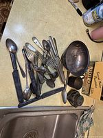 Photo showing a collection of tarnished vintage forks, spoons, knives, and hollowware including a large bowl and cup with heavy patina and wear on a kitchen countertop.