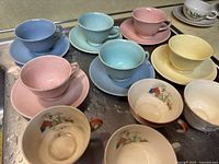 Various vintage teacups and saucers laid out on a counter showing pastel solids and scenic transferware cups.