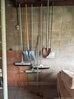 Photo showing full set of garden tools hung against a wall including brooms, shovels, rake, and hoe.