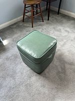 Front perspective of the green vinyl ottoman on carpet. Shows compact square shape, slight crease on top, side seams and side handles.