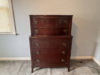 Front view of a tall mahogany highboy dresser with six drawers and brass oval hardware, showing subtle carved details on the top drawers and fluted trim.