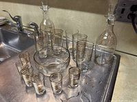 Photo showing the full set arranged near a sink with 2 tall decanters with stoppers, 6 tall glasses, 6 small glasses, 1 bowl, and 3 additional coordinating smaller glass pieces. All clear glass with horizontal gold bands.
