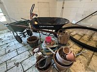 Overview photo showing black Craftsman wheelbarrow, green Scotts lawn spreader, red dolly, various clay and plastic pots, wooden basket, pink pail, plant trays, flexible hose, and plant care spray jug.