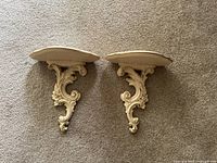 Top-down view of two ornate wall shelf sconces placed side by side on carpeted floor showing carved scroll and floral motifs, ivory and gold finish.