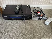 Panasonic 4-Head Omnivision VHS VCR on left with remote and AV cables, Philco DVD player on right with remote and AV cables, placed on carpeted floor.