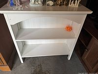 Front view of the small white wood bookshelf showing two shelves, arched apron, and minor surface wear.