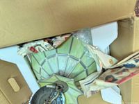 Top view of stained glass pendant light packed in box with newspaper wrapping and styrofoam for protection, showing green and white segmented glass panels with leaded framing and attached metal hanging chain with ceiling mount plate.