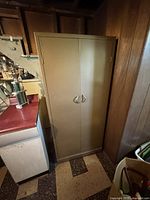 Front view of the vintage tall metal cabinet closed with dual doors and handles in a basement or garage setting.