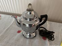 Front-angle view of shiny chrome electric coffee percolator showing the curved spout, black handle, decorative finial and cloth-wrapped cord.