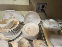 Photo showing stacked dinner plates, small plates, tea cups on saucers, and a covered tureen with lid, all decorated in delicate pink and yellow floral patterns with scalloped edges and black detailing.