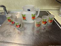 Seven pieces of frosted glassware on a metal surface. Centerpiece is a pitcher with a handle and pouring lip, decorated with hand-painted tomatoes and an original blue Gay Fad label. Surrounding the pitcher are six frosted tumblers with matching tomato graphics.