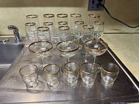 Front view of all 20 glassware pieces arranged on metal sink surface, showing height difference and gold rim details.