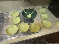 Apple-themed ceramic salad set: 1 large green apple bowl, 8 yellow apple bowls, 2 yellow serving utensils.