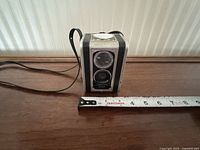 Front and side view of the Kodak Brownie Hawkeye camera, showing the twin-lens reflex design and wrist strap with measuring tape for scale.