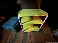 Vintage metal utility cart, three tiers with lime green and yellow trays, white metal frame with wheels, shown in dim light on floor