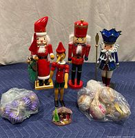 Four wooden Christmas figures including two nutcrackers in red and blue, a Pinocchio figure, and Santa Claus, shown with a small nativity scene and three bags of Christmas ball ornaments on blue fabric.