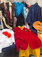 Wide shot showing multiple adult costumes including the red Robin vest with yellow pants and cape, white clown outfit with red pom-poms, and other hanging costumes in blue and brown.
