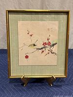 Front view of the framed Asian-inspired artwork showing the bird and red blossoms on a branch within a gold-tone bamboo-look metal frame.