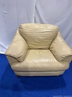 Front view of beige leather armchair with wood legs on blue protective mat and white backdrop, showing plush cushions and wide armrests.