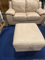Beige leather loveseat in background with matching ottoman in foreground on moving blanket
