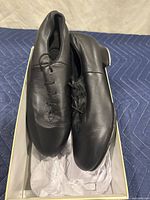 Top view of the pair of black leather tap shoes inside the original box showing lace-up closure and smooth leather surface.