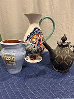 Three decorative ceramic pitchers with different hand-painted designs and colors displayed together on a blue fabric surface.