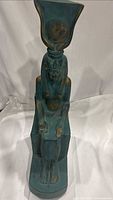 Photo shows front view of the tall seated Egyptian goddess statue with turquoise-green paint and gold accents on the headdress and body.