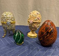 Set of four decorative egg-shaped trinket boxes on blue quilted surface showing varied colors, detailing, and sizes