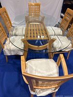 Full view of glass dining table with six light wood chairs arranged around it, showing glass tabletop with light scratches and chair cushions with fabric covers.