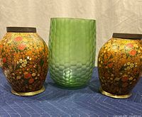 Three vases displayed on a blue padded surface: one green honeycomb glass vase in the center, flanked by two vintage floral vases with metallic bases and dark rims.