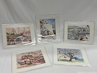 Five small giclee art prints arranged on white surface, all sealed in plastic. Subject matter includes marinas and coastal town views.