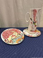 Photo showing ceramic pitcher on small plate and oval platter with colorful nature designs including a peacock.