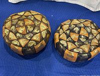 Two Moroccan leather pouf ottomans viewed from above on a blue protective fabric, showing circular multi-colored leather patchwork with Egyptian and animal motifs in tan, black, brown, and gold tones.